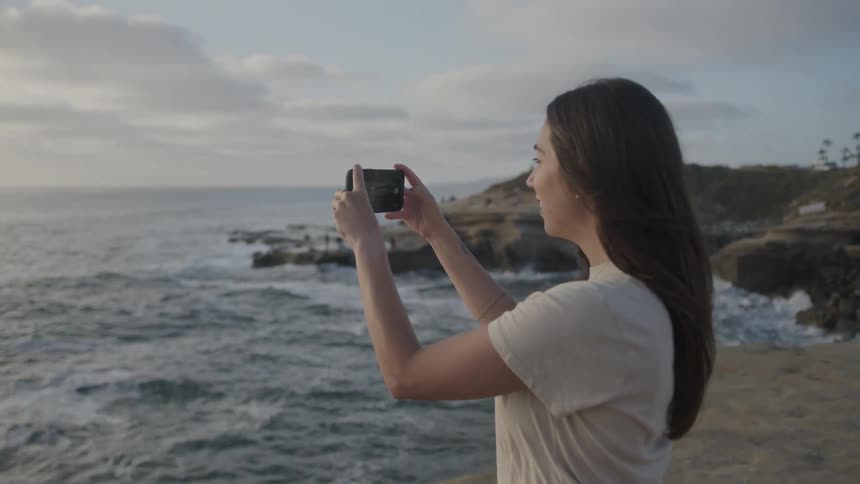 A young woman standing on a cliff using her smartphone to film while smiling