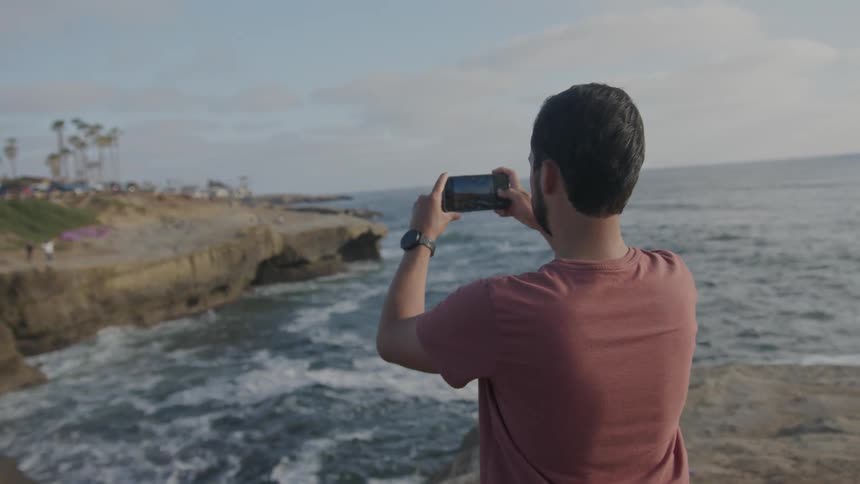 A man takes a photograph of a coastal area with a smartphone