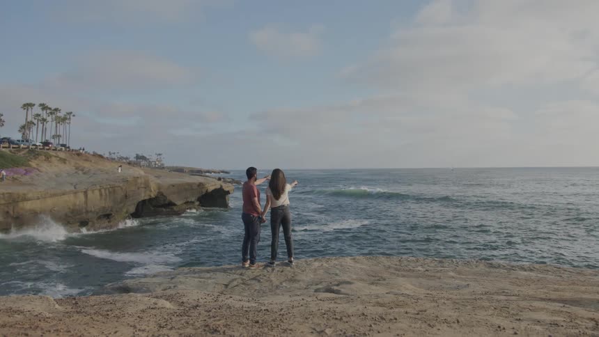 View of a couple holding hands back to back walking on a rock by the sea