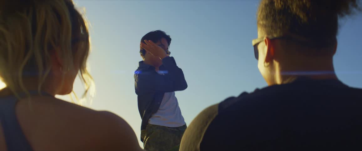 A man and a woman both dressed in black tops and wearing sunglasses watch as a dancer performs with sunlight in the background