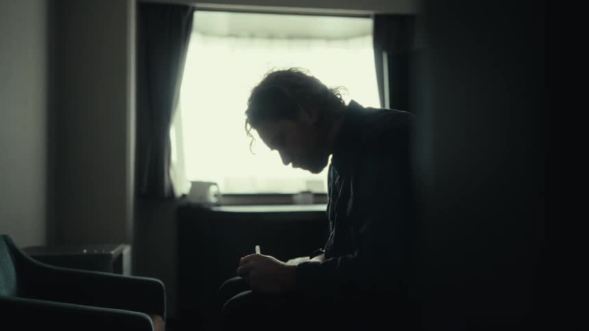 A bearded man wearing a dark shirt sits on the bed writing in a notebook in a room