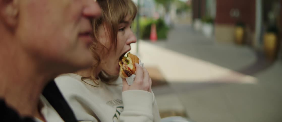 A woman in a white sweater eats her sandwich while talking to a man sitting on a bench in the street