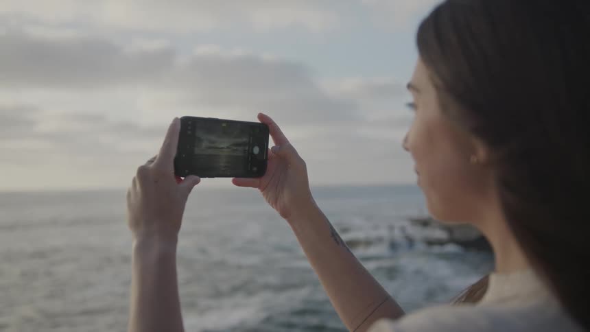 Woman on a beach photographing the sky and sea with her iphone