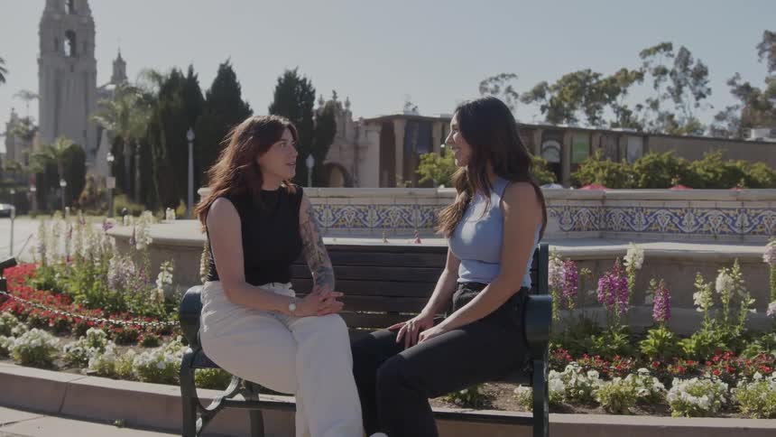 Two women sit on a bench on a college campus and talk