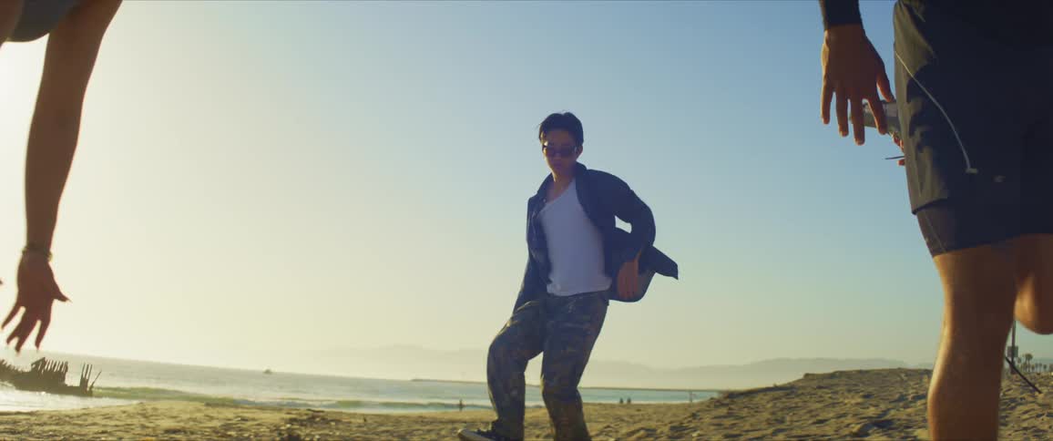 Two people stretching their legs and in the background a man dancing on the beach sand