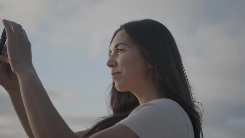A woman takes pictures of the ocean with her smartphone at dusk