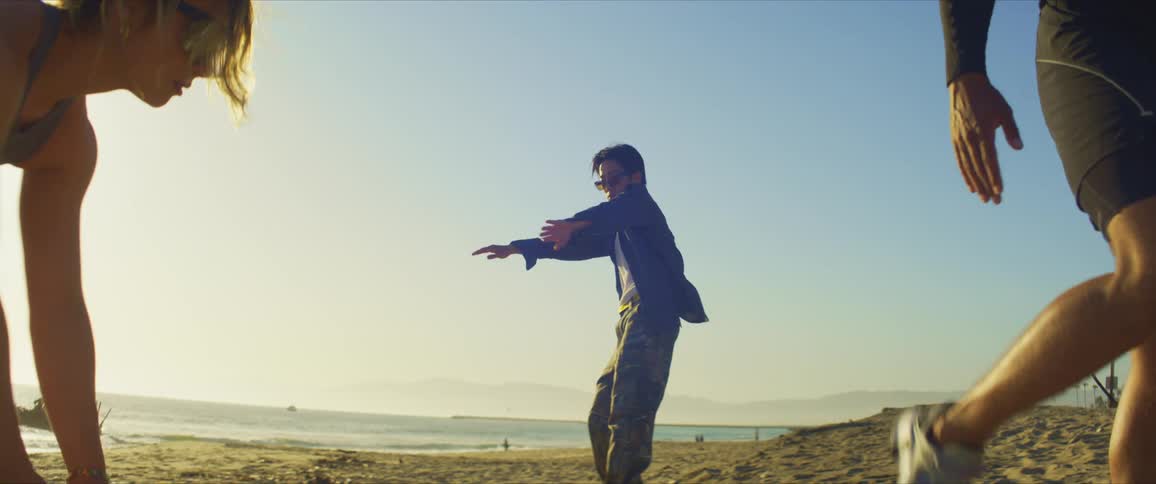Man and woman in sports outfits warm up while a dancer dances in the background on the beach at sunset