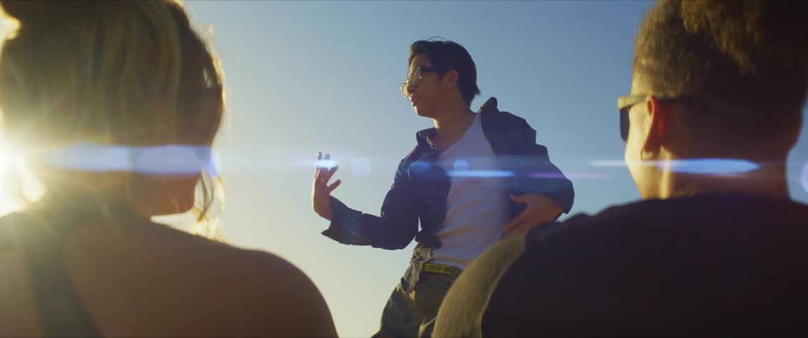 A man and a woman both wearing sunglasses earrings and black tops watch a male dancer wearing sunglasses a blue shirt and white pants on the beach at sunset