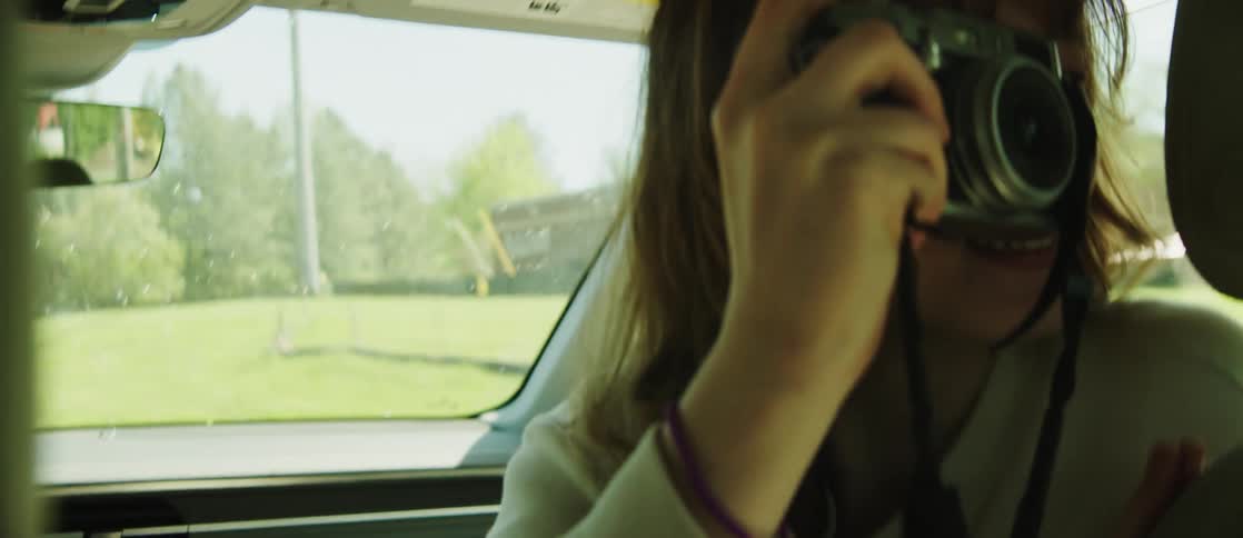 A woman with wavy hair and a white shirt sits in the front seat taking a photo with a camera