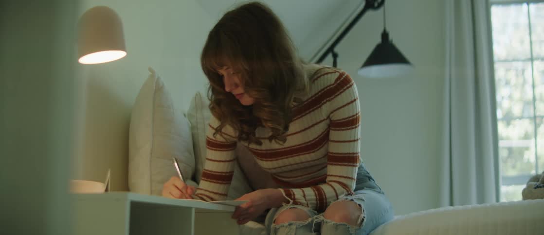 Woman sitting on the bed and writing on a paper on the dresser next to her