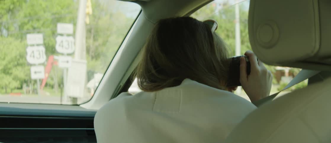 A woman with wavy hair and a white shirt sits in the front seat taking a photo with a camera while the car is driving
