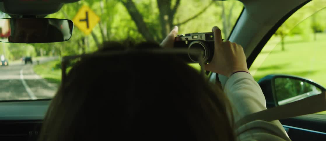 A woman with wavy hair and a white shirt sits in the front seat taking a selfie with a camera while the car is driving