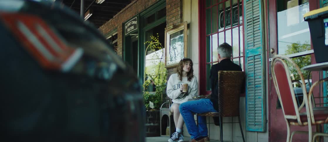 A man and a woman sit outside a quaint caf engaging in conversation while she holds a cup of coffee