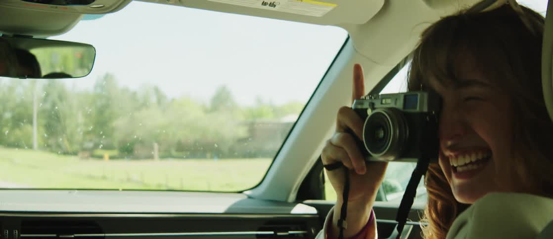A woman with wavy hair and a white sweater sits in the front seat of a moving car taking photos of the driver laughing and enjoying the moment
