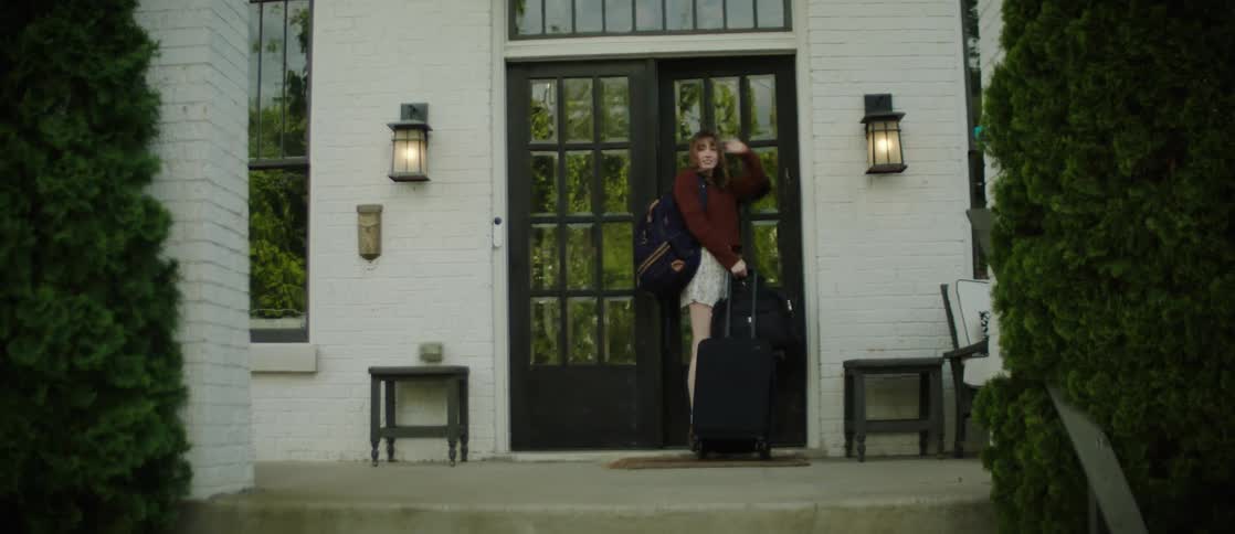 Young woman at the entrance of a house with a suitcase waving goodbye