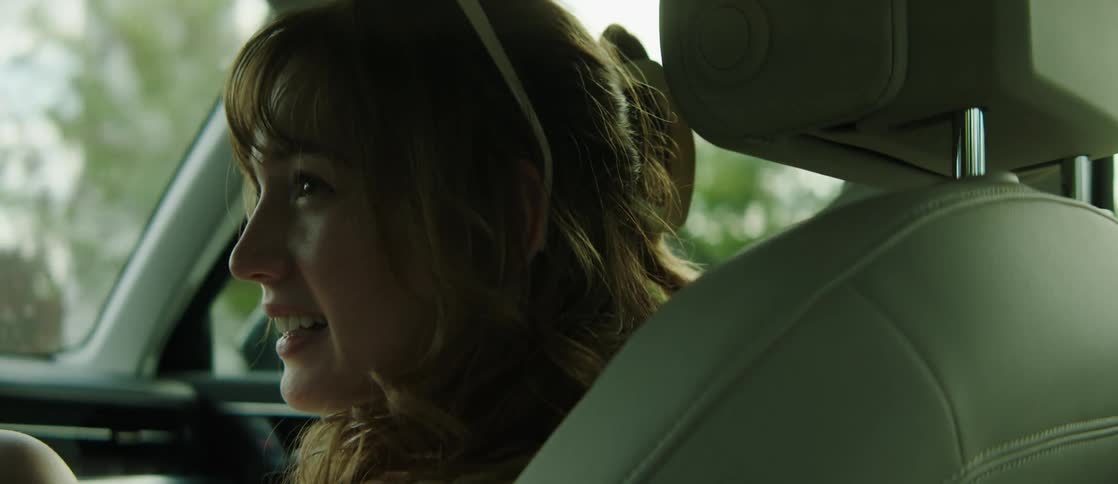 A woman with brown wavy hair and white sunglasses sits in the front seat of a moving car talking with the driver