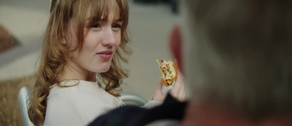 A middleaged man talks with a woman with brown wavy hair eating a sandwich while sitting on a metal bench