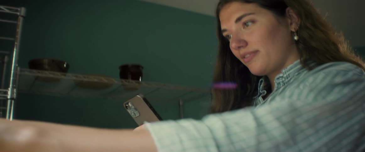 Woman with brown wavy hair earrings and a plaid shirt takes photos with an iphone smartphone in the kitchen