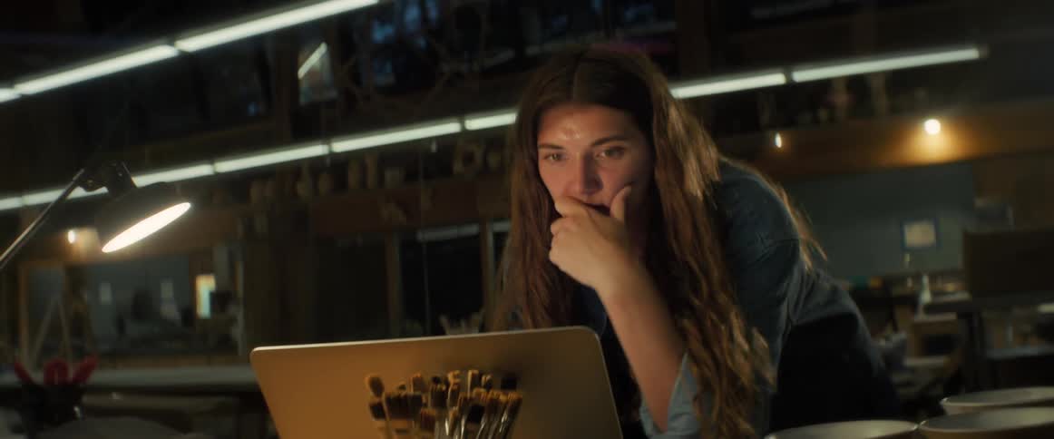 A woman with brown wavy hair and a blue shirt work on laptop in a workshop