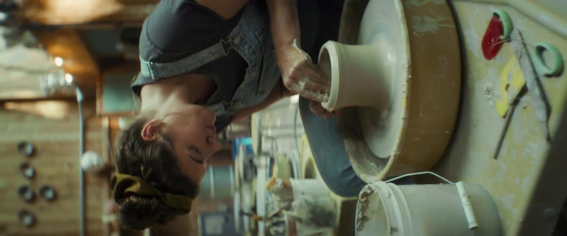 A woman in a denim jumpsuit shaping a lump of clay on a pottery wheel