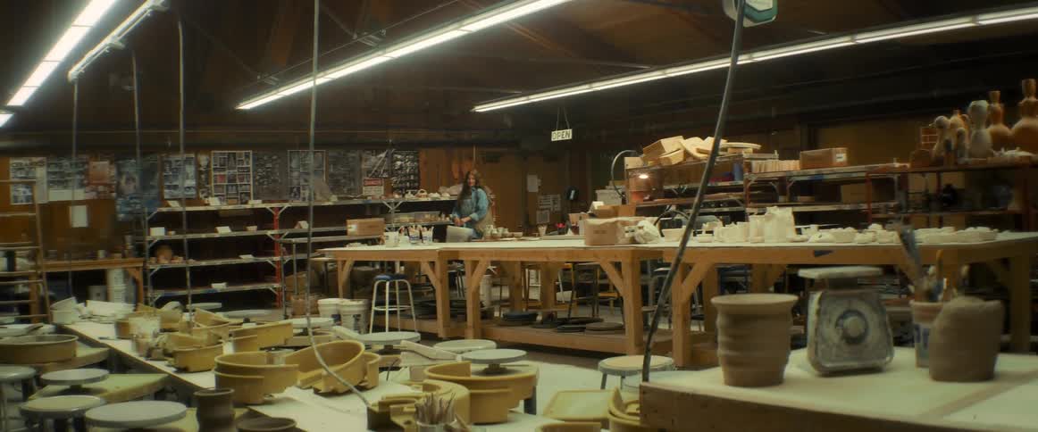 A woman wearing a blue shirt and jumpsuit turns on the lights and enters the pottery workshop