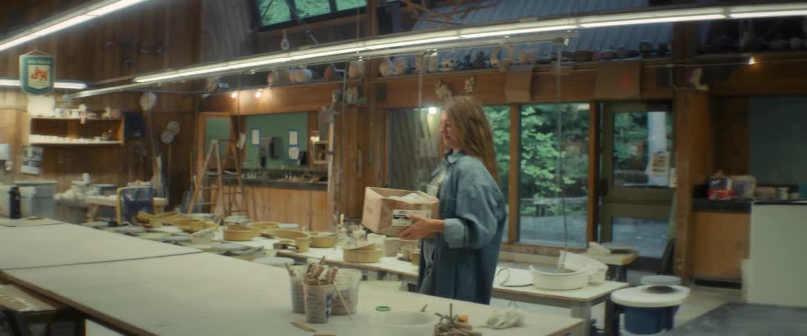 Woman with brown hair and a blue shirt carries a box in a pottery workshop