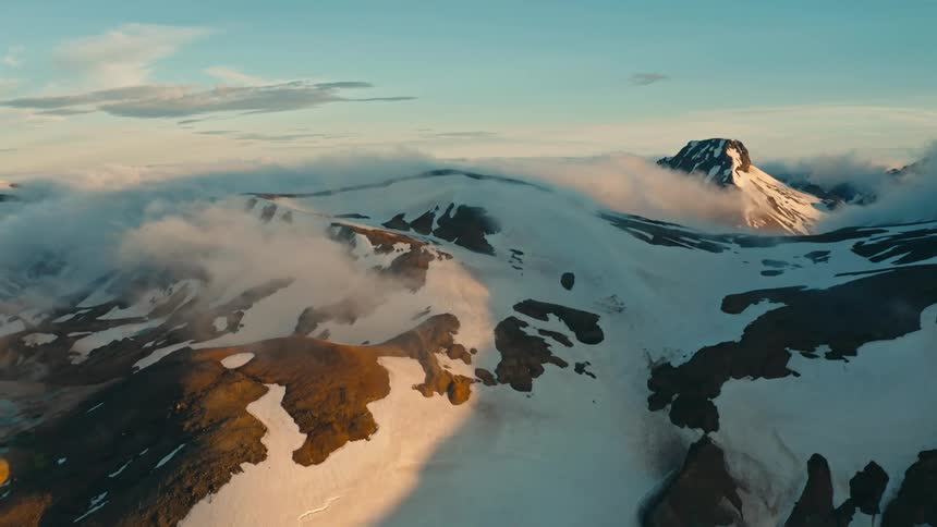 An aerial view of a mountainous landscape with patches of snow and clouds hovering around the peaks