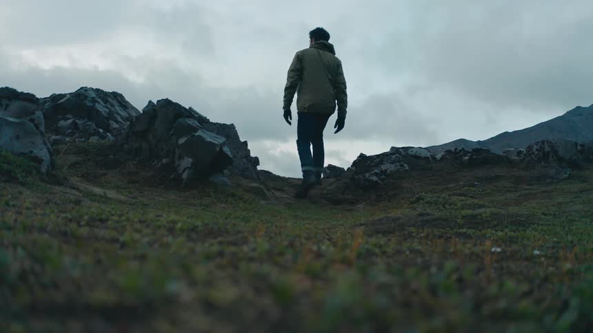 A man wearing a jacket and pants walks up to the mountain top to watch the scene on a cloudy day