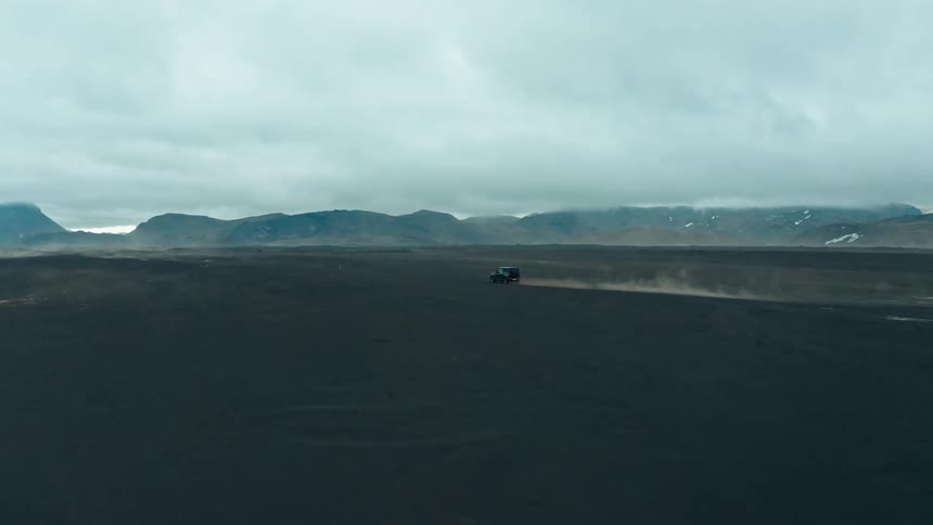 Black car speeding through volcanic soil with mountains around on a cloudy day