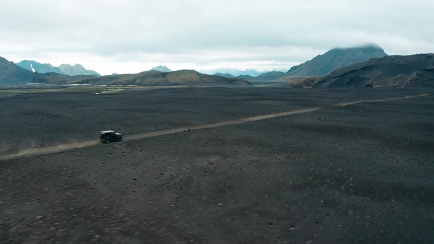 Aerial panorama of a black vehicle traveling along a dirt road with mountains around it on a cloudy day