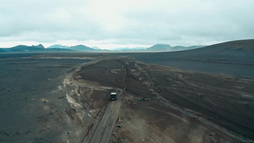 Aerial scenery of a black vehicle traveling on a black ground with mountains around it on a cloudy day