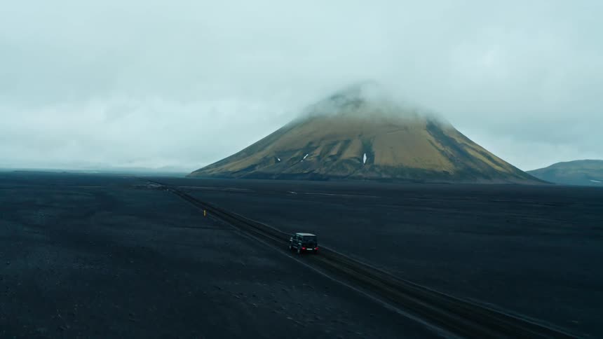 Aerial shot of a black car driving on black ground with a volcano in the background on a cloudy day