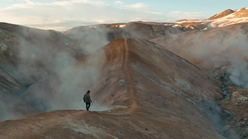 A person walking on a ridge overlooking a geothermal area with steam rising from the ground