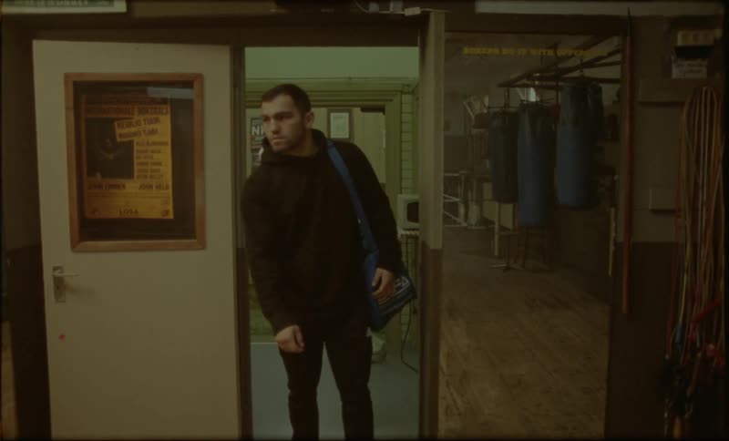 A man wearing a black hoodie pants white shoes and a blue shoulder bag enters the boxing gym turns on the lights and looks around