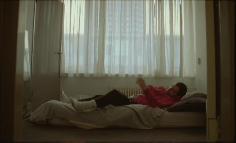 A man lying in a mattress on the floor of a small room forces himself to get up
