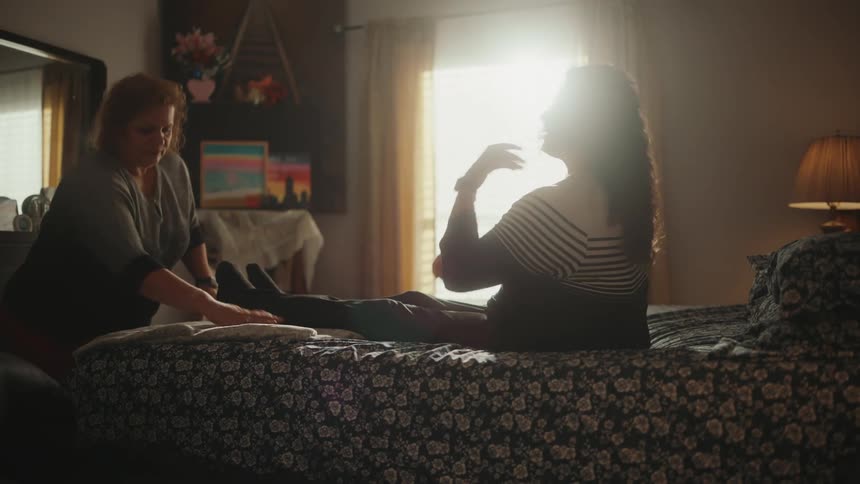 A home aide assists a handicapped woman in laying down on the bed