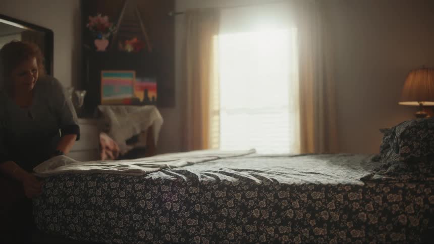 Home aide straightening the bed in a room illuminated by sunlight