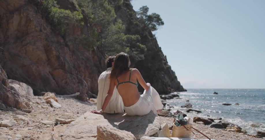 Two individuals seated on a rocky beach facing the ocean on a sunny day