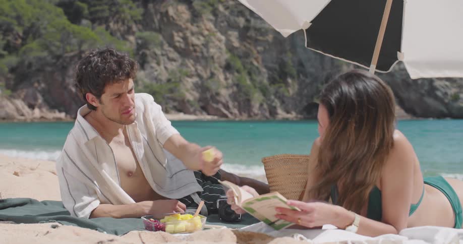 Man eating fruit and giving it to his wife who is next to him reading a book