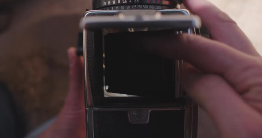 Mans hands holding a vintage camera photographing a woman lying on the sand reading a book and then closing the camera