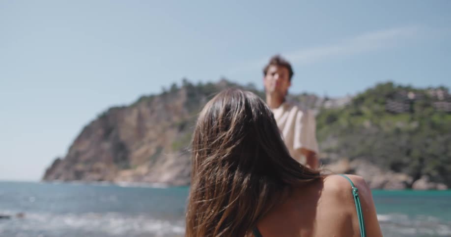 A man wearing a white shirt and blue striped shorts stands in front of a woman in a bikini who is sitting on the rocky beach at the bay
