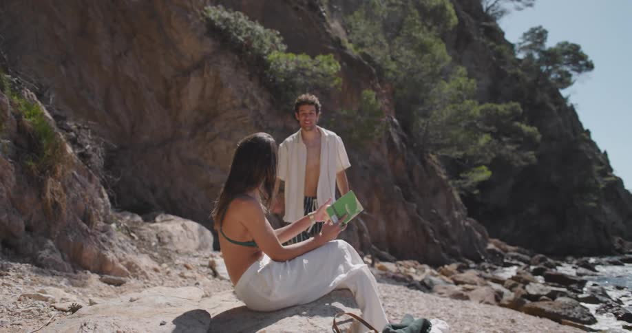 A young man joins a woman who is reading a book on the beach