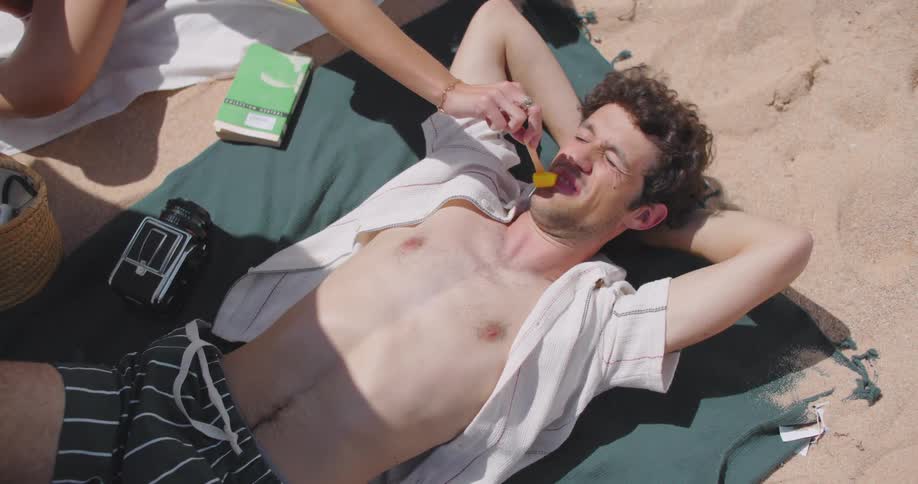 Man lying down relaxing on the beach sand and a womans hand placing a slice of fruit in his mouth