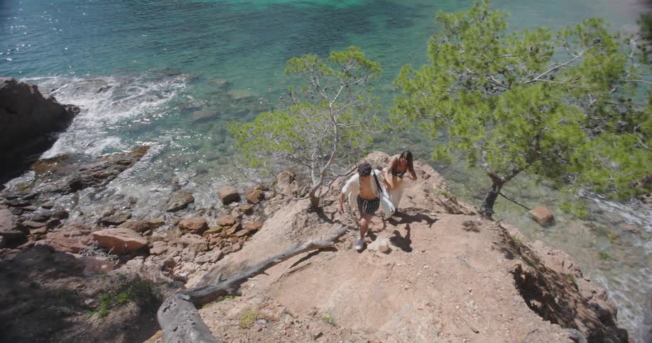 Man holding hands with his wife helping her climb a hill on the edge of a turquoise beach