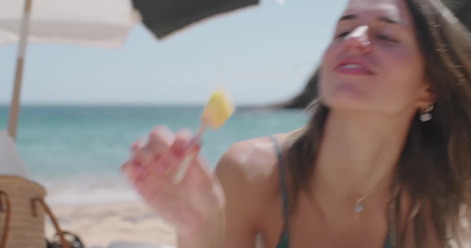 Woman lying on the sand towel on the beach eating a fruit and reading a book