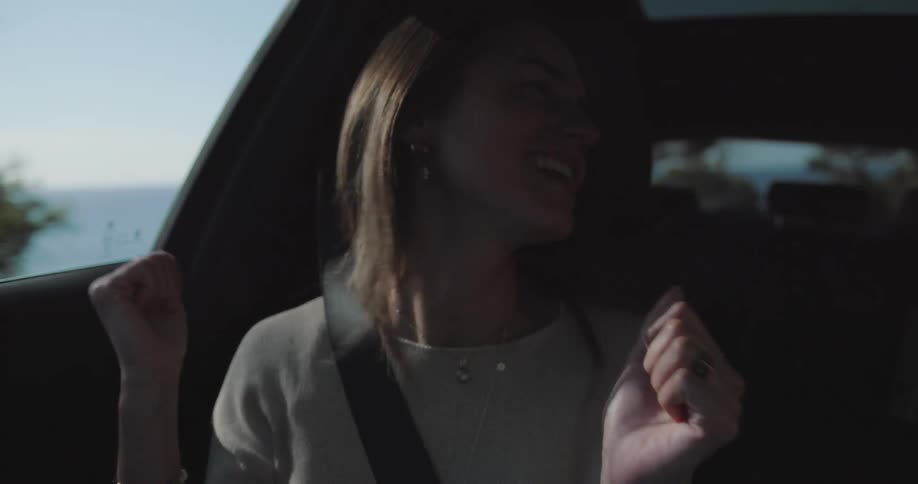 A young happy woman laughs and smiles while dancing in a car driving near the ocean