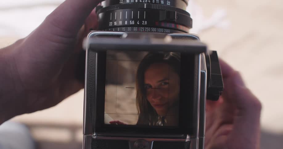 Closeup of hands holding a vintage camera with the viewfinder framing a woman reading a book at the beach