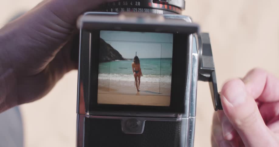 Hands holding a vintage camera with a woman on a beach seen in the viewfinder