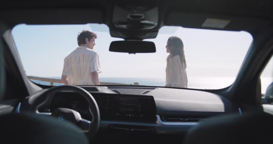 A young couple sitting on the front of a car next to the ocean
