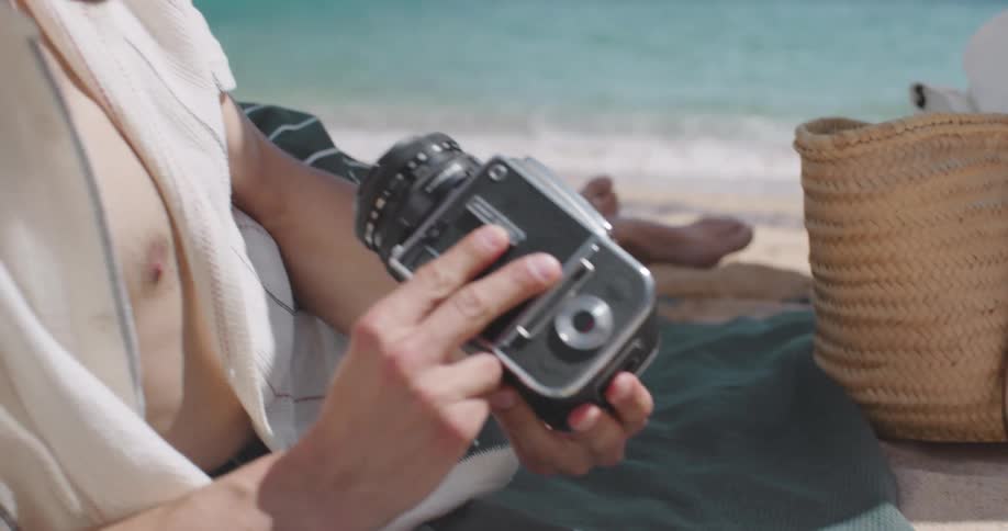 A young man handles a vintage camera next to a woman reading a book in the beach
